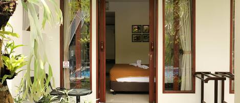 A hotel room with a bed, a small table, and a chair. There are plants outside the room.