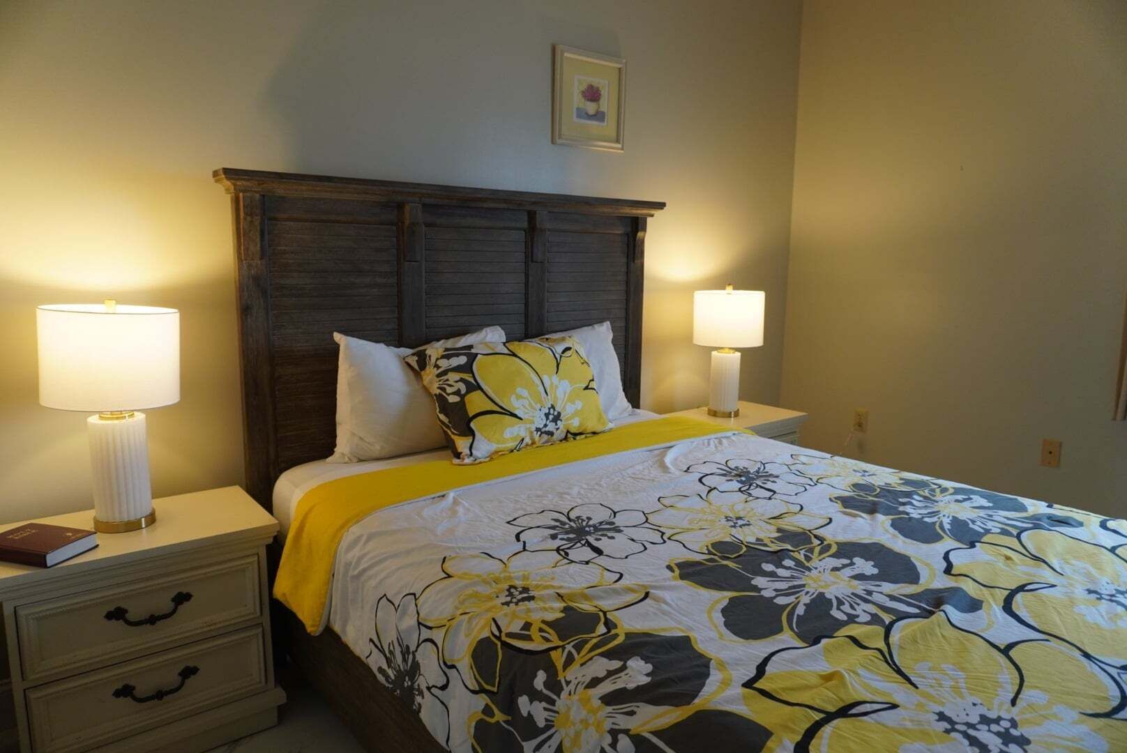 Een net opgemaakt bed met een bloemenbedekking, twee nachtlampjes, een nachtkastje met lades en een ingelijst beeld aan de muur.