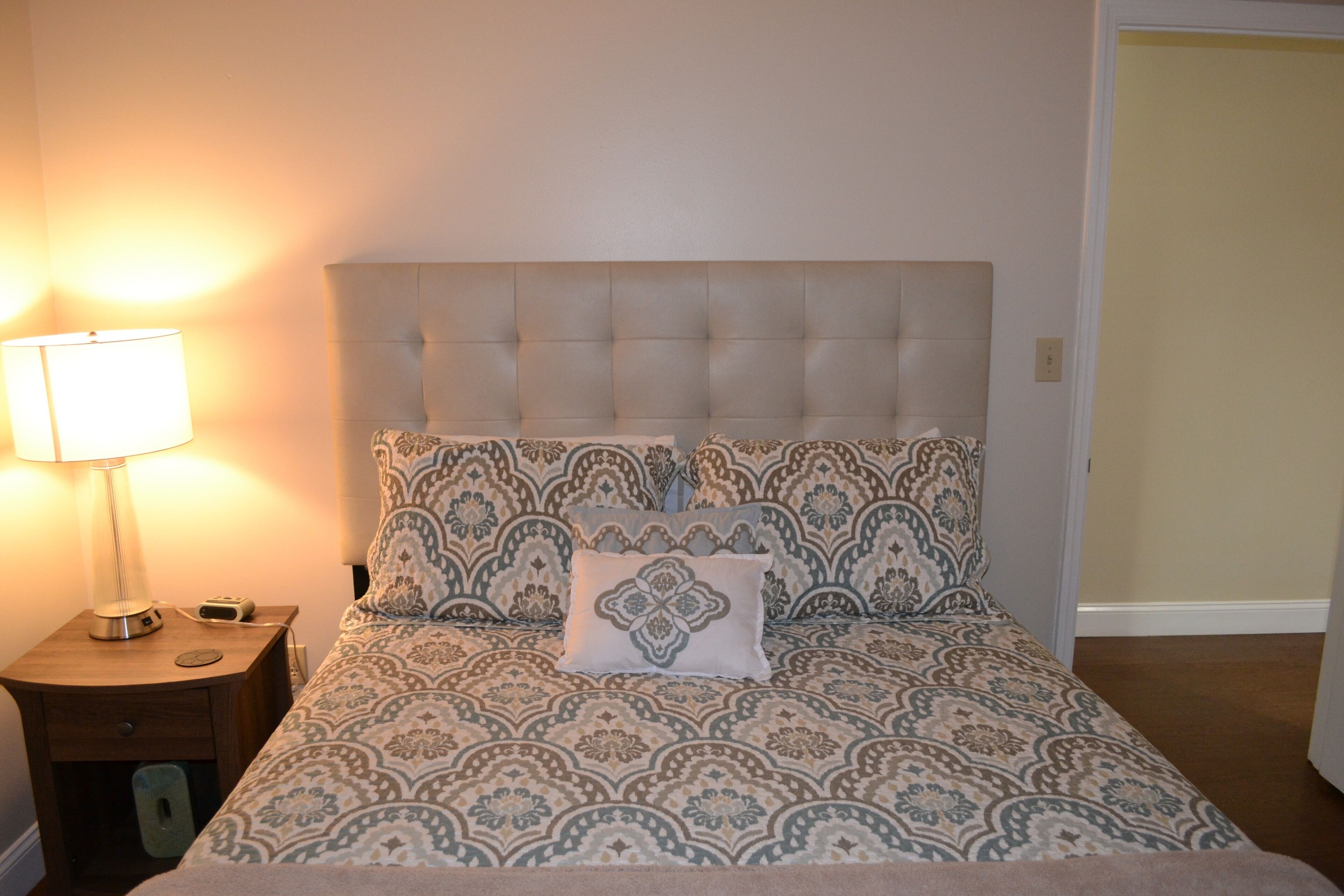 A bedroom with a tufted headboard, patterned bedding, a bedside lamp, and a nightstand with a lamp and a phone.