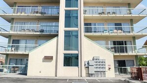 A multi-story building with balconies, a central vertical feature, and a parking area with a handicapped parking sign.