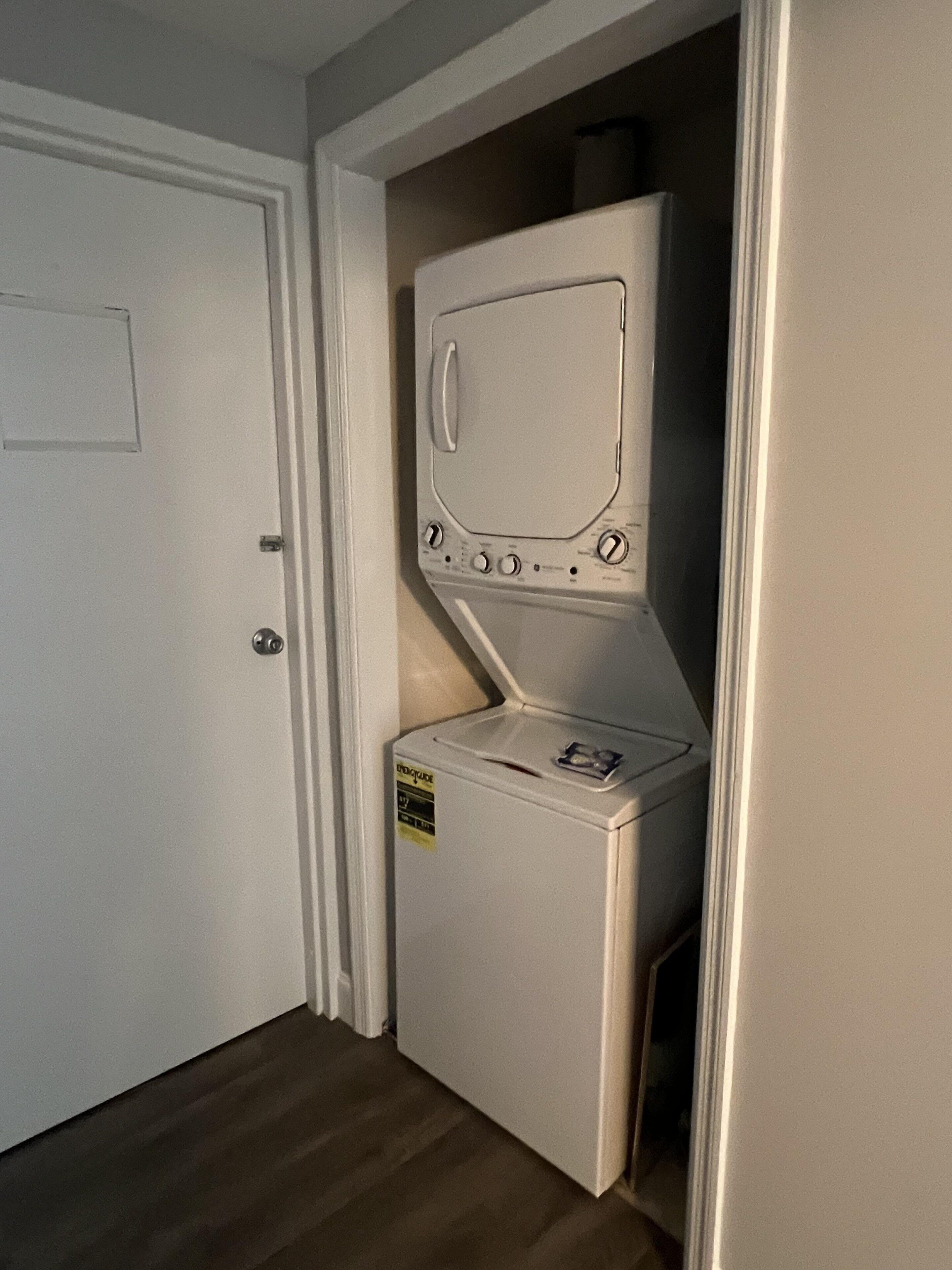 Eine kompakte, weiße Waschmaschine in einem kleinen Raum mit Holzboden und einer weißen Tür.
