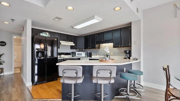 A modern kitchen with dark cabinets, a black refrigerator, a microwave, and a coffee maker. There are two bar stools and a wooden table.