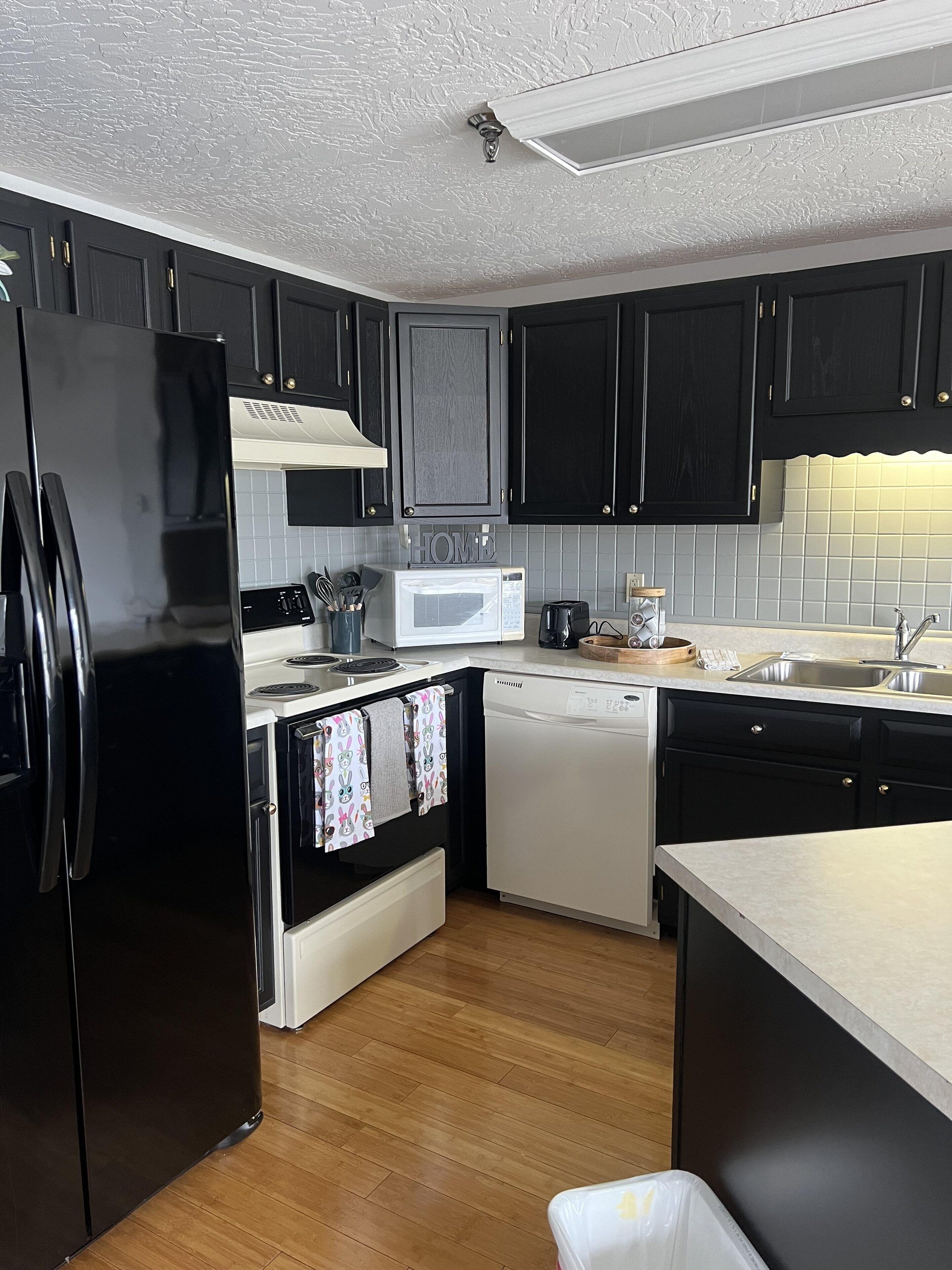 A modern kitchen with dark cabinets, a black refrigerator, a microwave, a dishwasher, and a wooden floor.