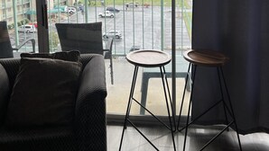 A room with a large window, a balcony with a table and chairs, and a view of a parking lot and buildings.