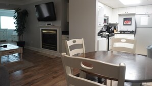 Ein Essbereich mit einem Holztisch und weißen Stühlen, einem Fernseher über einem Kamin und einer Küche mit weißen Schränken und Geräten.
