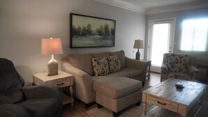 Ein Wohnzimmer mit einer Couch, einem Hocker, einem Couchtisch, zwei Lampen, einem Gemälde und einem gemusterten Sessel.