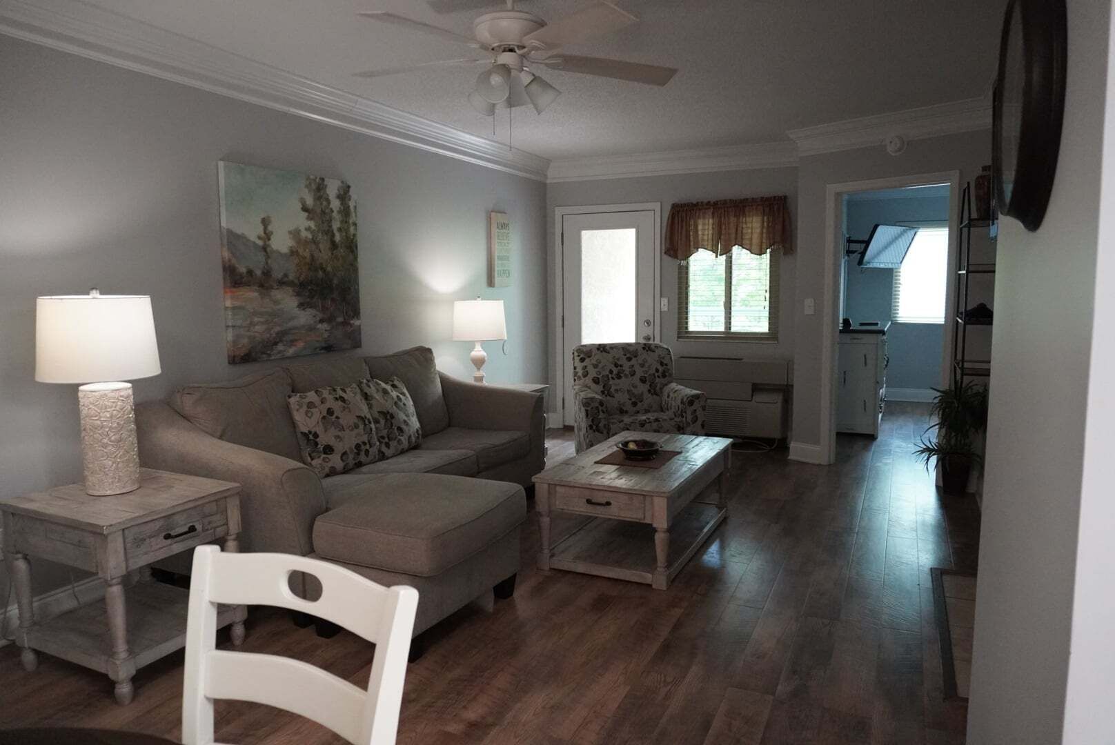 A living room with a sofa, armchair, coffee table, and a white chair. There is a ceiling fan, a painting on the wall, and a window with curtains.