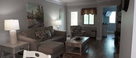 A living room with a sofa, armchair, coffee table, and a white chair. There is a ceiling fan, a painting on the wall, and a window with curtains.