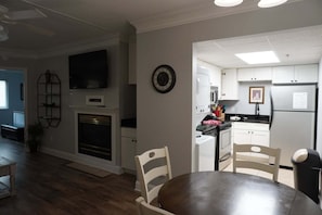 A compact kitchen with white cabinets, a microwave, a stove, and a refrigerator. There is a dining table with chairs, a television mounted on the wall, and a clock on the wall.