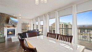 Ein geräumiges Wohnzimmer mit Kamin, eine Essecke mit großem Tisch und ein Balkon mit Blick auf ein Gebäude und Berge.