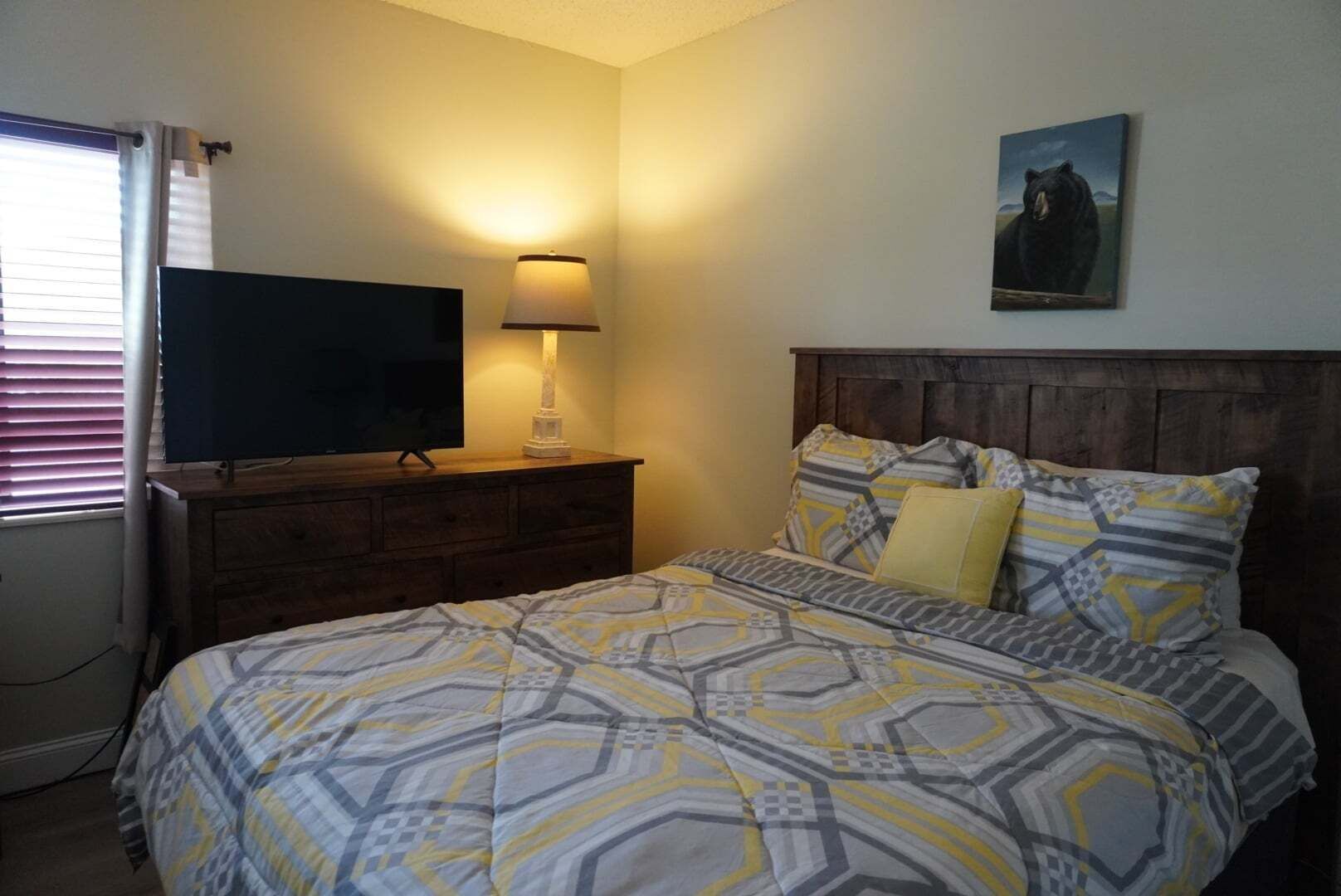 A bedroom with a bed, a TV on a wooden dresser, a lamp, and a bear painting on the wall.