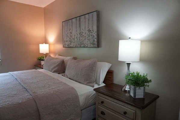 A neatly arranged hotel room with a bed, two bedside lamps, a small table with a plant, and a wall-mounted artwork.