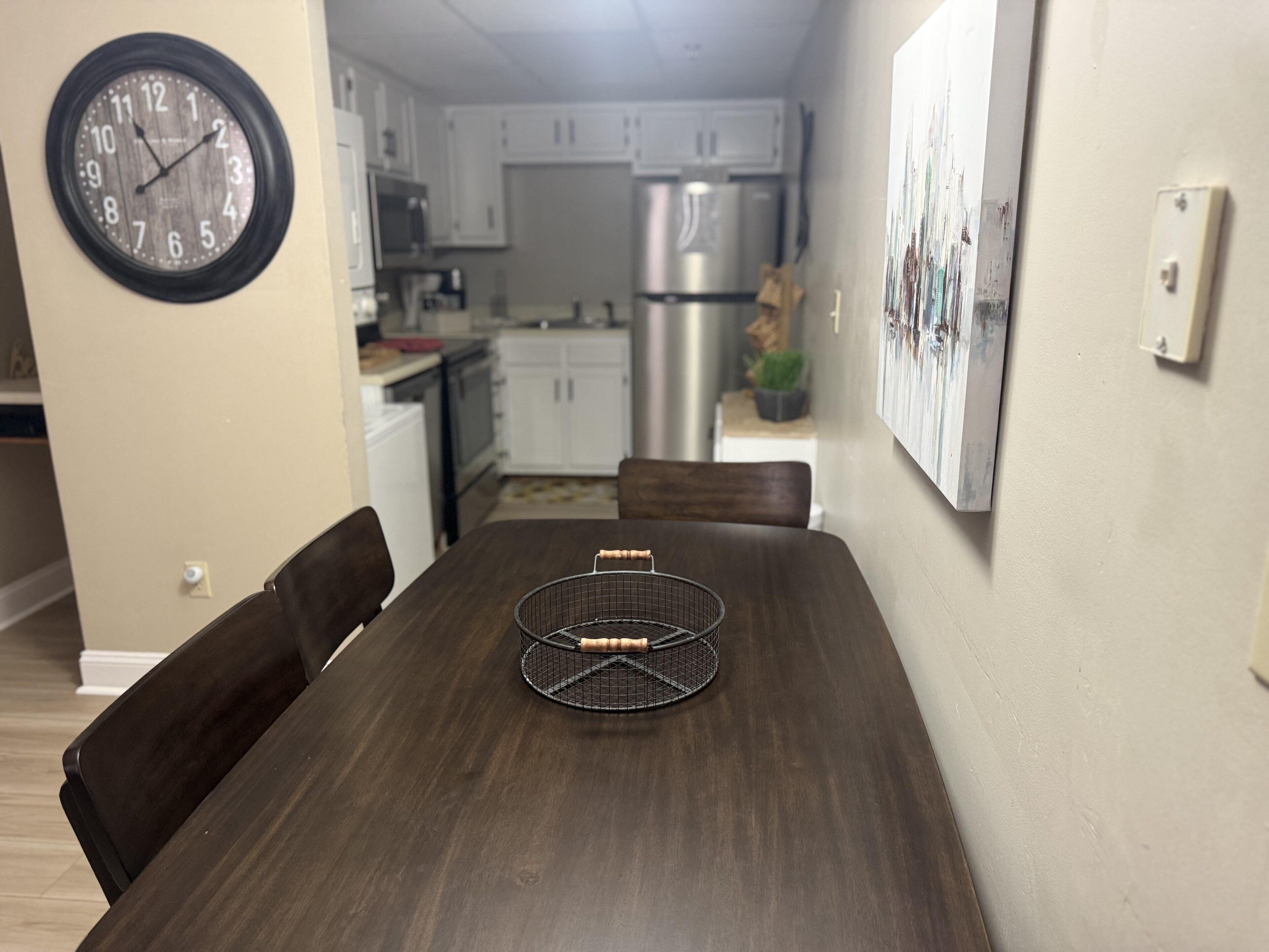 A dining area with a wooden table, chairs, a clock, and a kitchen in the background.