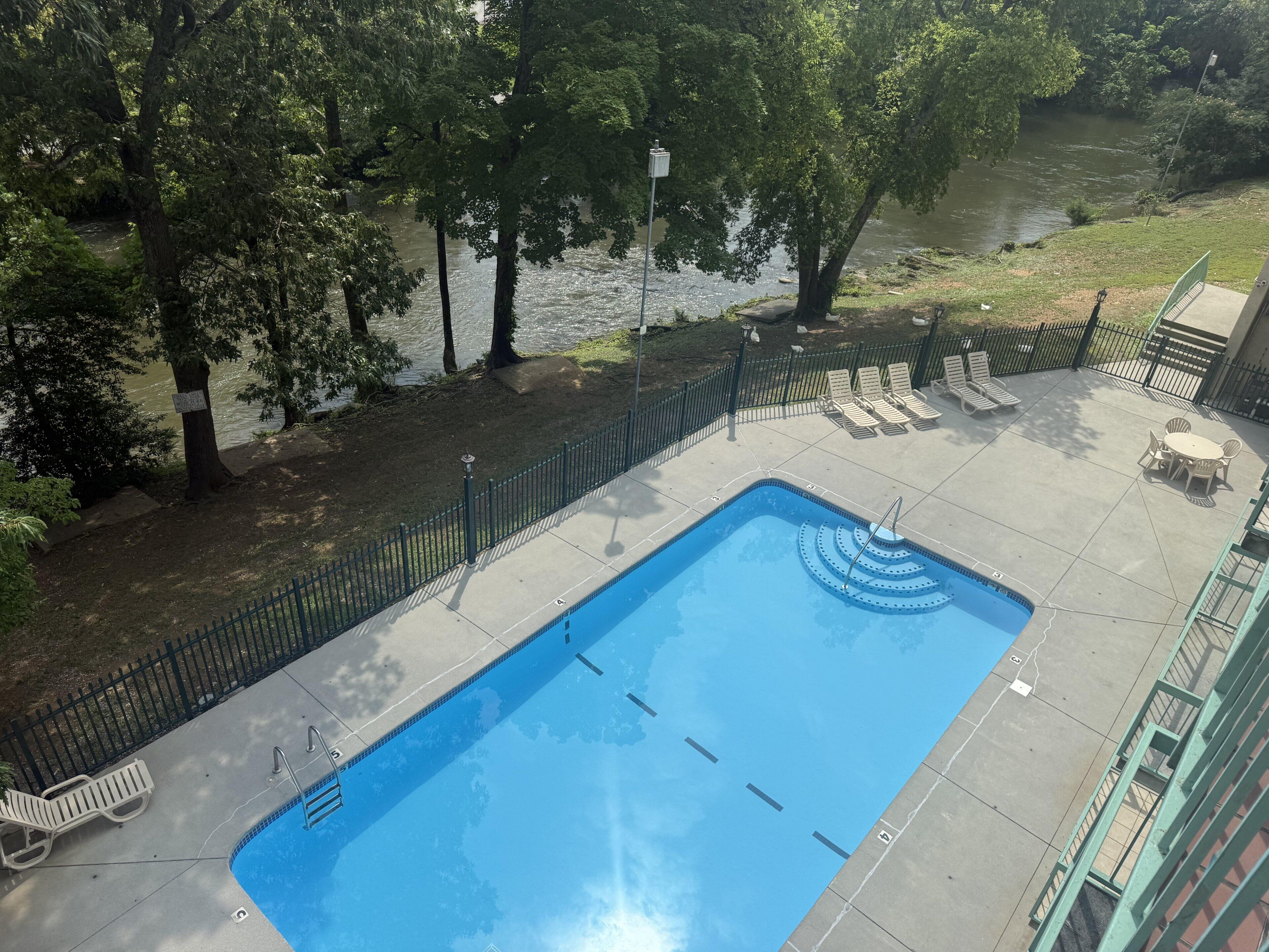 Piscina rectangular al aire libre con una escalera metálica, rodeada por una cerca verde, y contigua a un río.
