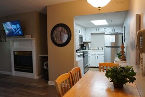 A dining area with a wooden table and chairs, a clock on the wall, a television displaying a game, and a kitchen in the background.