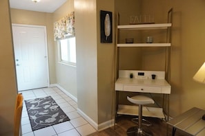 A hallway with a white door, a window with floral curtains, a white desk with a chair, and a shelf with decorative items.