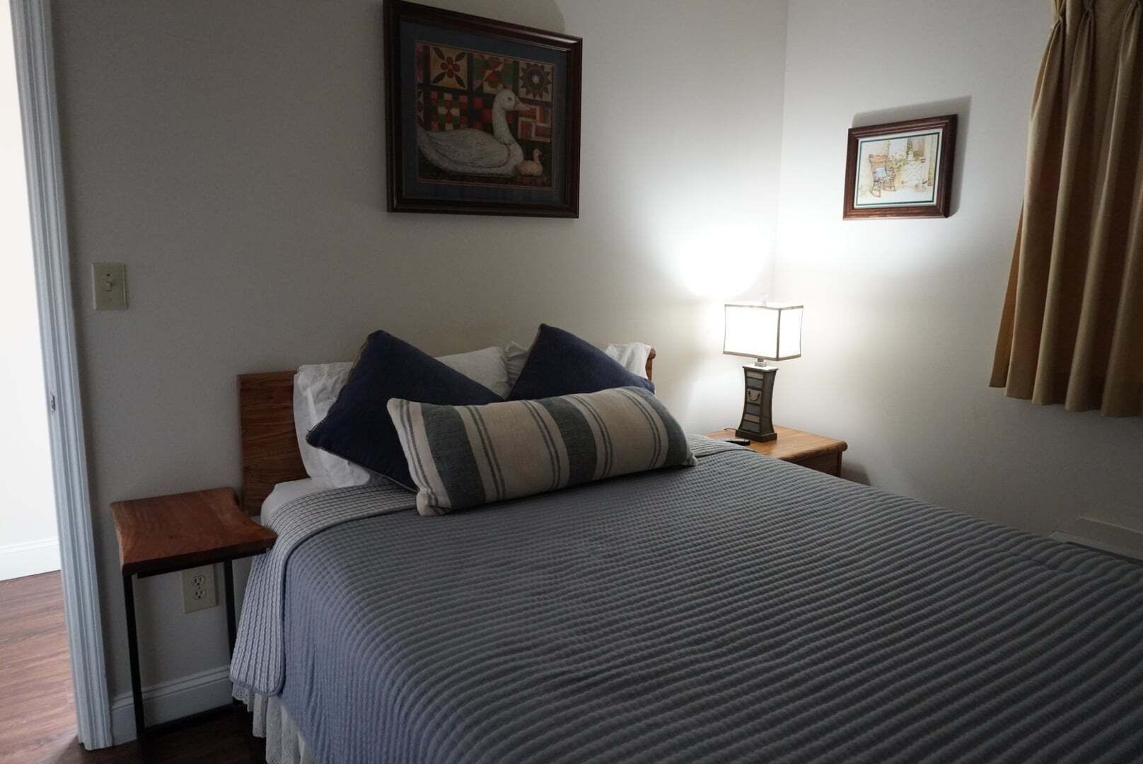 A neatly made bed with a striped bedspread, two pillows, a nightstand with a lamp, and two framed pictures on the wall.