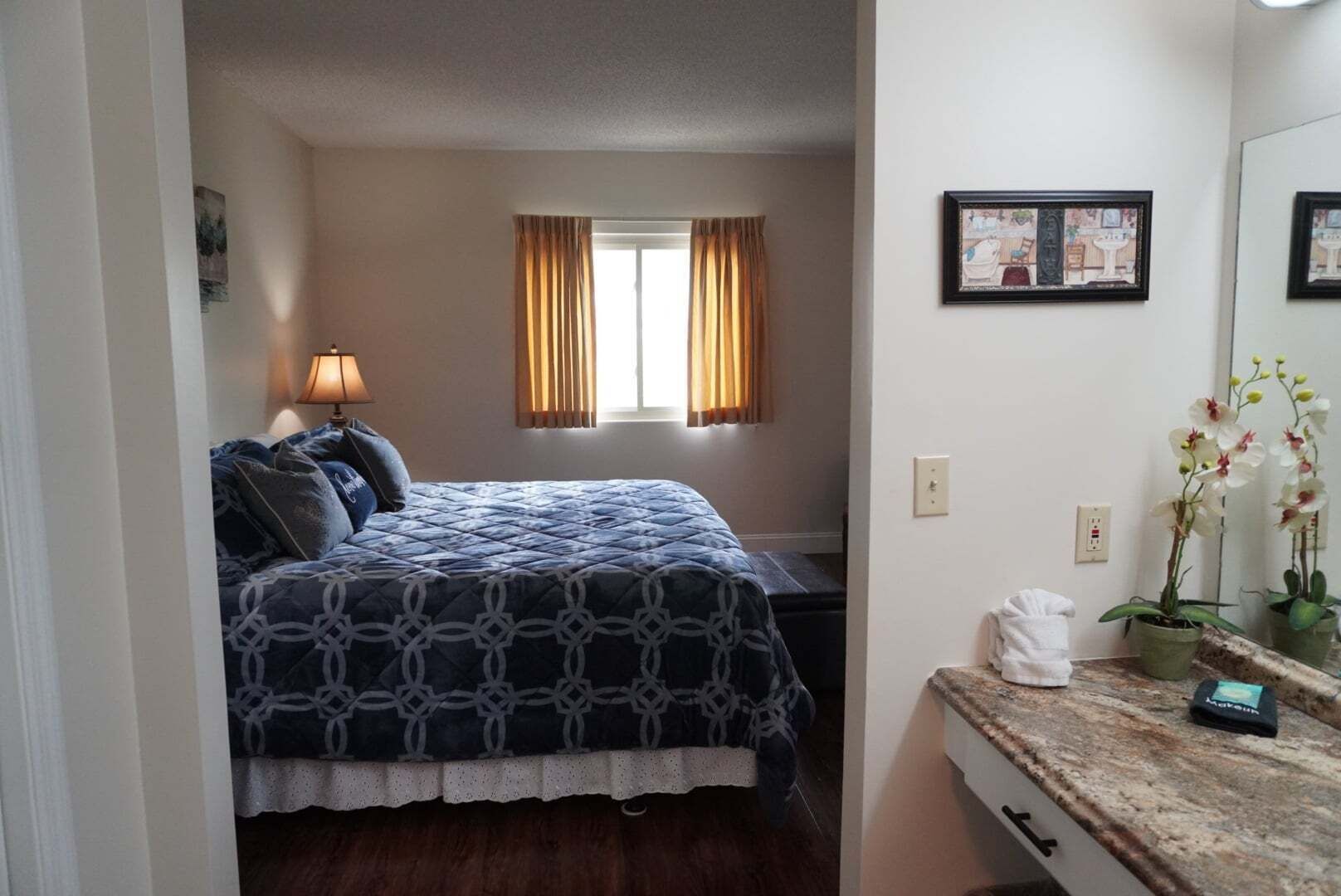 A bedroom with a bed, a nightstand, a lamp, a window with curtains, a mirror, and a small potted orchid.