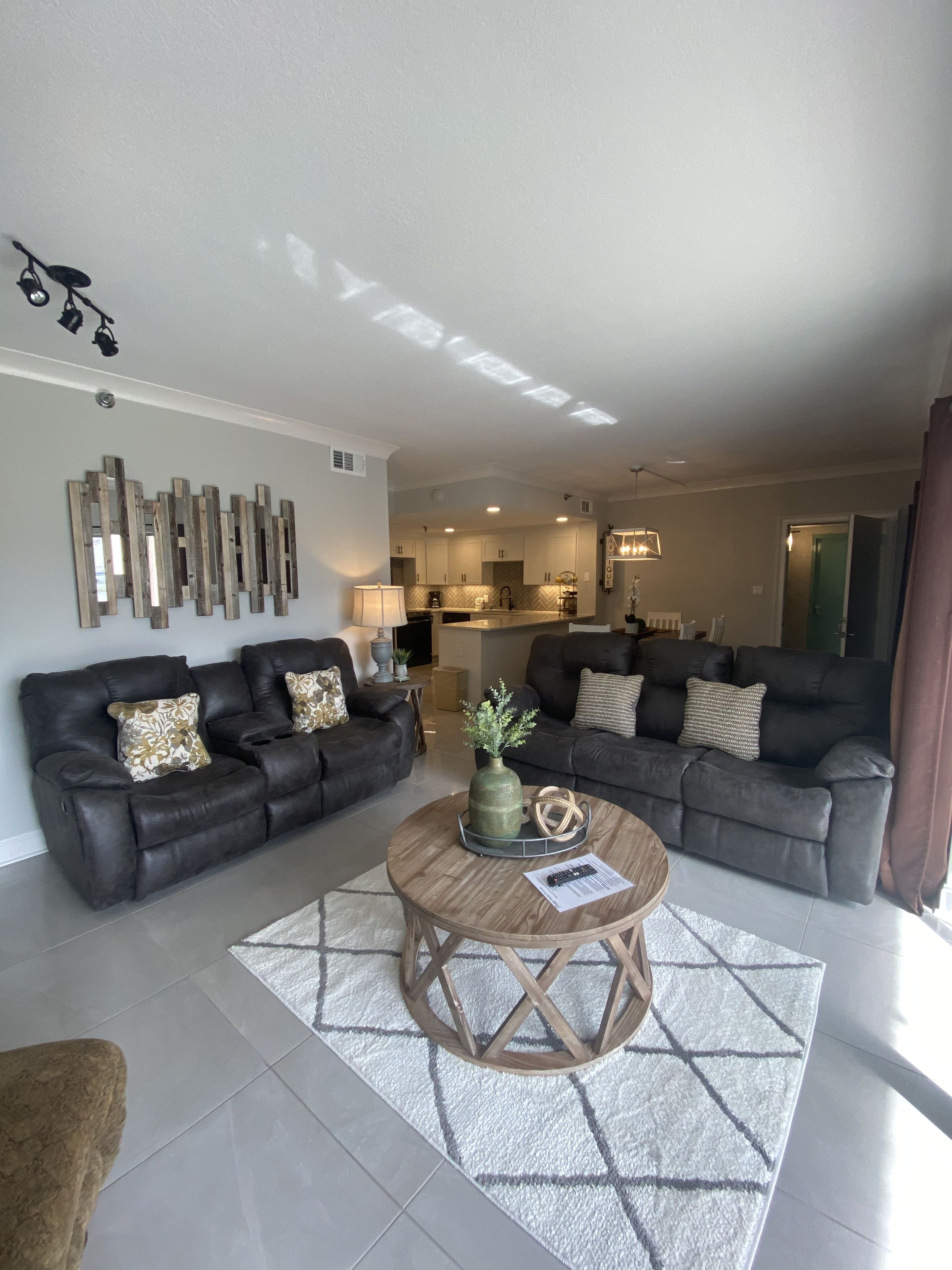 A modern living room with two dark leather sofas, a wooden coffee table with decorative items, a kitchen in the background, and a large mirror on the wall.