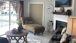 A living room with a sofa, armchair, and a fireplace. There is a large window with blinds, a clock, and a television on the wall. - 111 3bd 3 Bedroom Condo by RedAwning (Pigeon Forge)