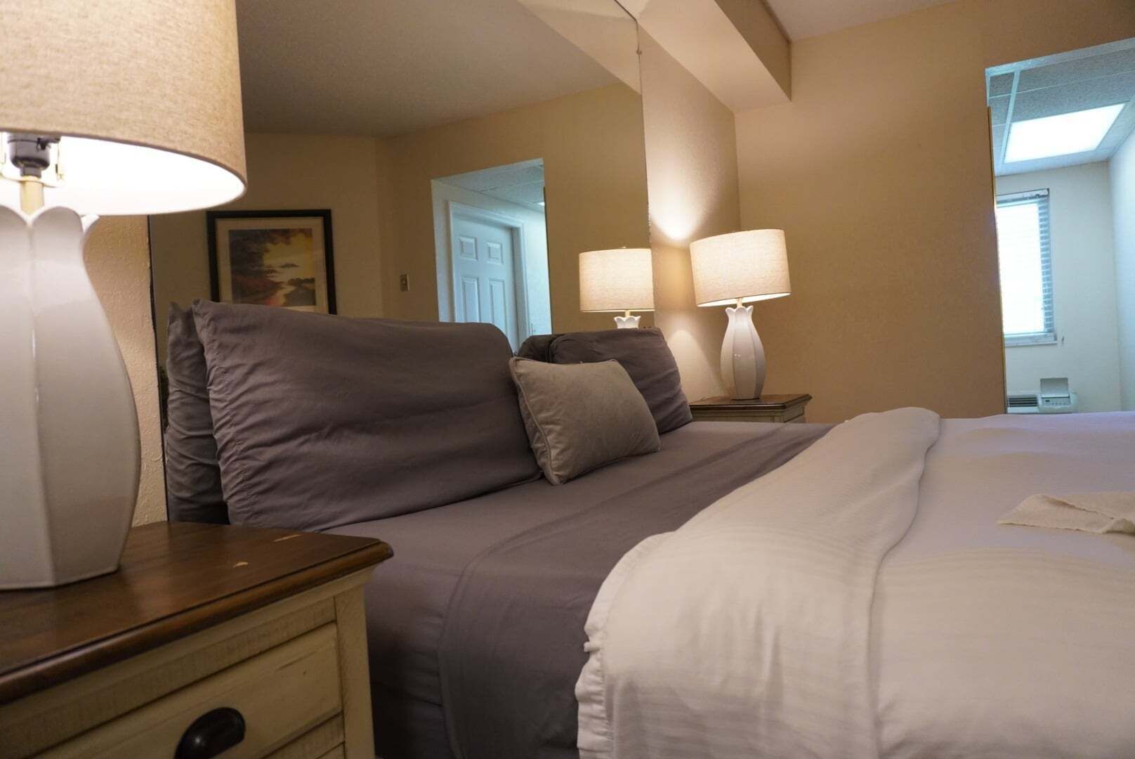 A hotel room with a bed, two bedside lamps, a nightstand, a wooden dresser, and a door leading to another room.