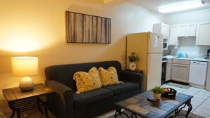 Ein gemütlicher Wohnbereich mit Sofa, Couchtisch, Lampe, einem Bild an der Wand, einer Küchenzeile mit Schränken und einem Kühlschrank.