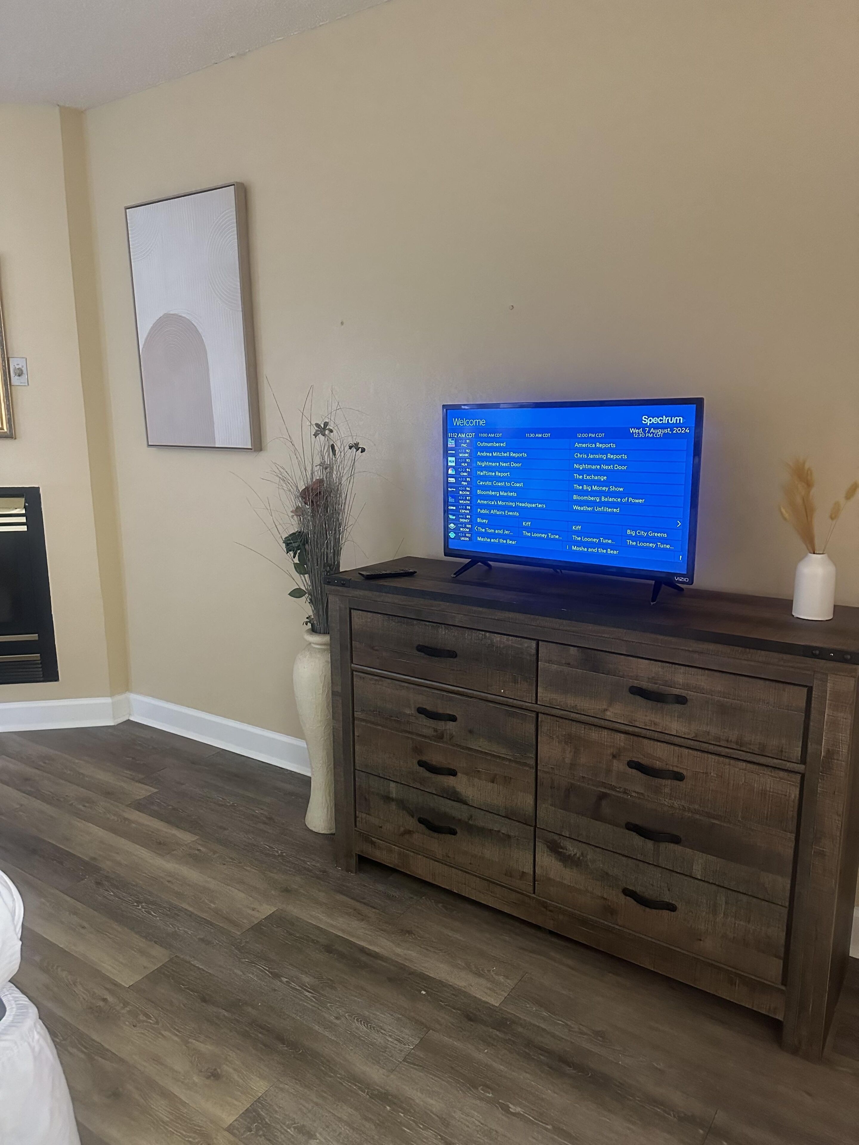 A living room with a television on a wooden cabinet, a vase with dried flowers, and a framed picture on the wall.