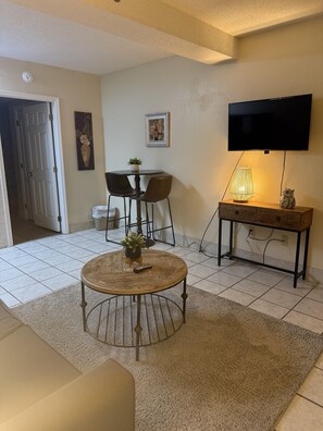 A living room with a round coffee table, a TV mounted on the wall, a dining area with chairs, and a small table with a plant.