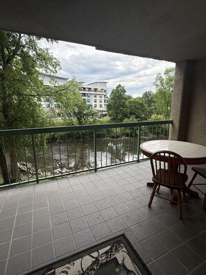 A balcony with a view of a river, a dining table with chairs, and a building in the background. - Riverside 209 1BR Condo with Jacuzzi by RedAwning (Pigeon Forge)