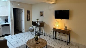 Ein Wohnzimmer mit rundem Couchtisch, einem an der Wand montierten Fernseher, einem Essbereich mit Stühlen und einer Küchenzeile inklusive Schränken und Geschirrspüler.