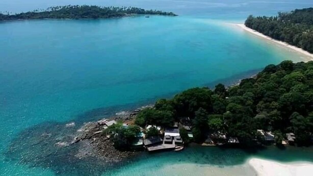 An aerial view of a coastal area with a beach, a small island with dense vegetation, and a few buildings near the shore.