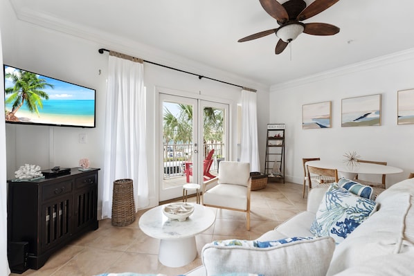 Un salon avec un téléviseur diffusant une scène de plage, un ventilateur de plafond, un canapé blanc avec des coussins bleus, une table à manger et une vue sur la plage à travers les portes-fenêtres coulissantes.