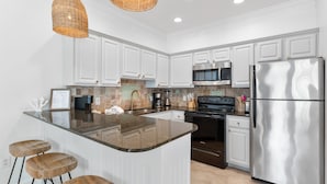 A modern kitchen with white cabinetry, a stainless steel refrigerator, a microwave, and a stove. There are three wooden stools at the island. - Crystal Beach. Caribbean Dunes. Completely Renovated. Corner Unit. Gulf View.  (Destin)