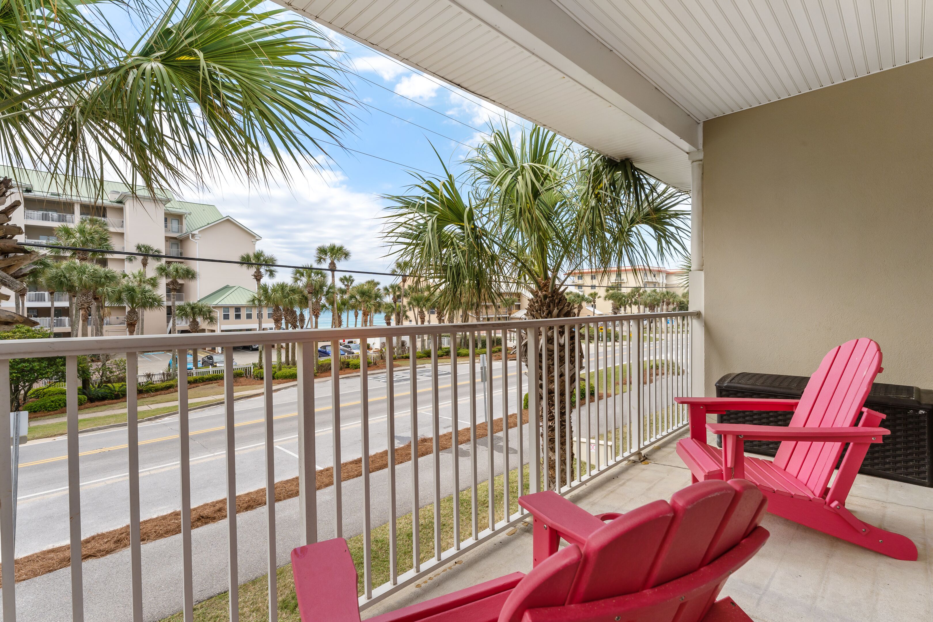 A balcony with a view of a road and palm trees, featuring two red Adirondack chairs and a small table.