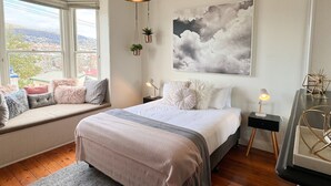 Ein Schlafzimmer mit Bett, Nachttischen, einer Sitzbank unter dem Fenster mit Kissen, einer hängenden Pflanze und einem großen Wandbild zum Thema Wolken.