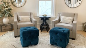 A living room with two beige armchairs, a small round table, and a blue ottoman. There is a Christmas tree with lights, a large mirror on the wall, and a window with blinds. - Spacious home w/Hot Tub near Lake Superior walk to restaurants, breweries, beach (Marquette)