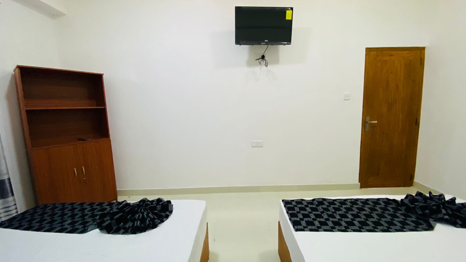 A room with two beds, a wooden wardrobe, a television mounted on the wall, and a door.