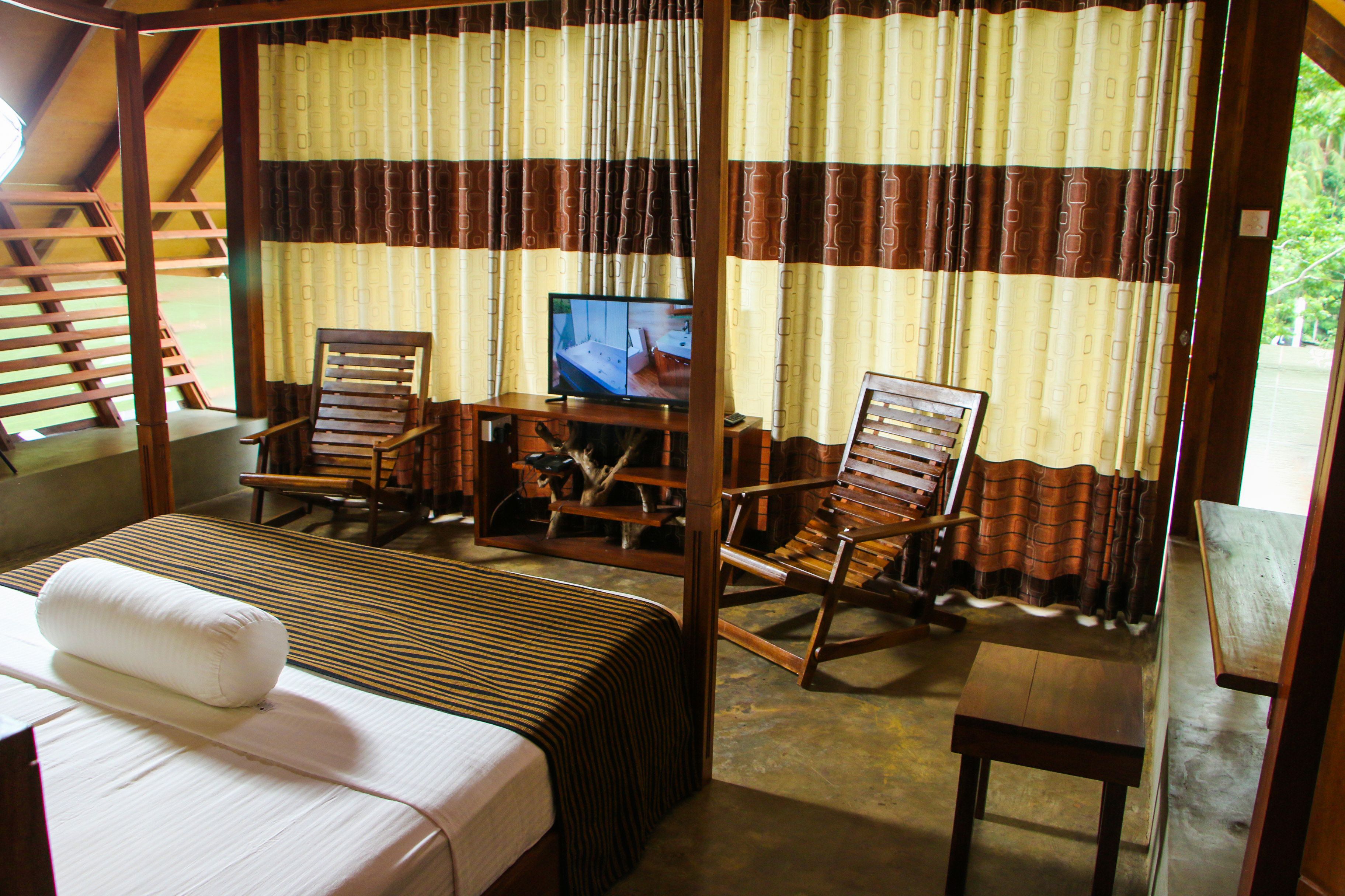 A room with a bed, two wooden chairs, a small table, a television, and curtains.