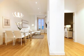 A modern living room with a glass dining table, white chairs, a sofa, and a coffee table. There is a chandelier, a framed picture on the wall, and a door leading to another room.