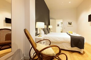 A hotel room with a bed, a chair, a nightstand with lamps, a mirror, and a television on the wall.