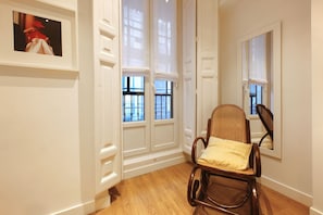 A room with a wooden floor, white walls, a wicker chair with a cushion, and a framed picture on the wall. - Chueca Exclusive (Madrid)