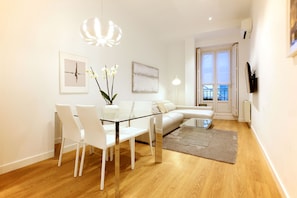 A modern living room with a glass dining table, white chairs, a white sofa, and a view of the balcony through the open door. - Chueca Exclusive (Madrid)