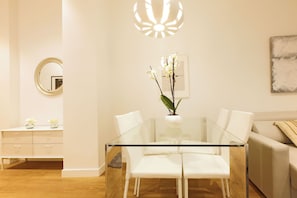 Un comedor moderno con una mesa de vidrio, sillas blancas, un florero con flores, un espejo redondo y una lámpara fijada a la pared.