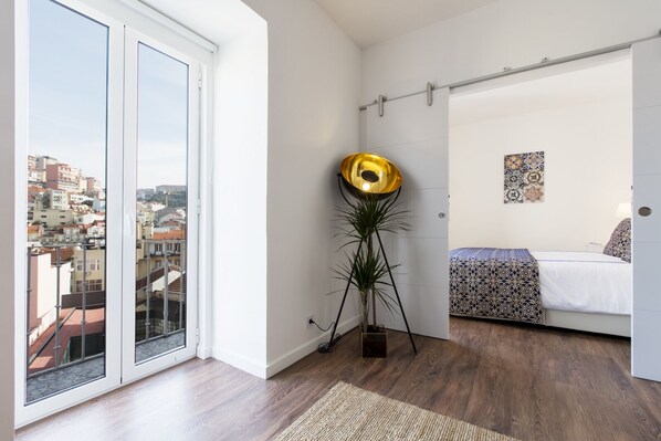 Een moderne slaapkamer met een groot schuifglasraam dat uitkijkt op een stadsgezicht, een vloerlamp, een bed met beddengoed met een patroon, en een klein kunstwerk aan de muur.
