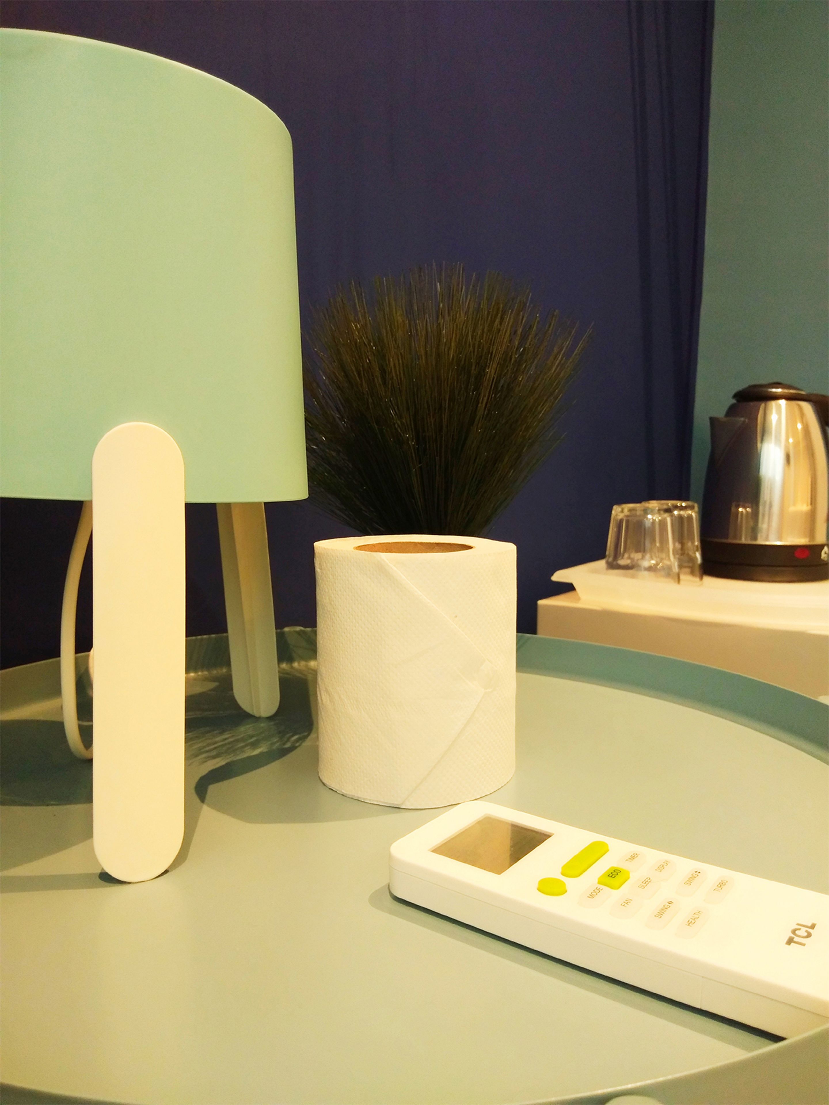 A bathroom with a toilet paper roll, a tissue box, a remote control, and a coffee maker on a table.