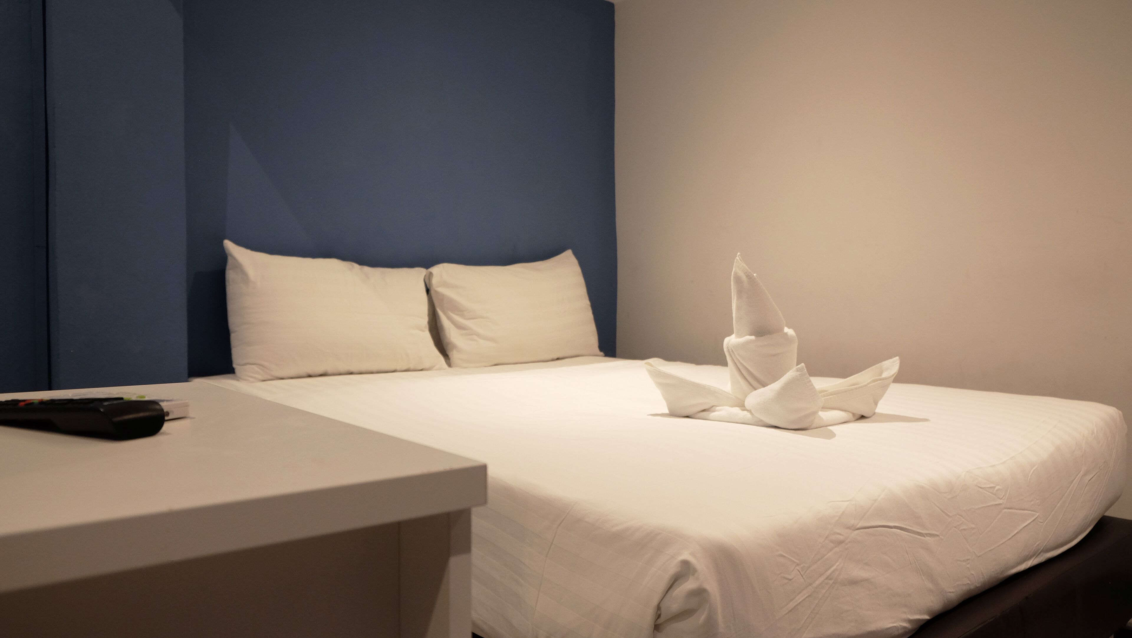 A hotel room with a neatly made bed, a remote control on the nightstand, and a blue accent wall.