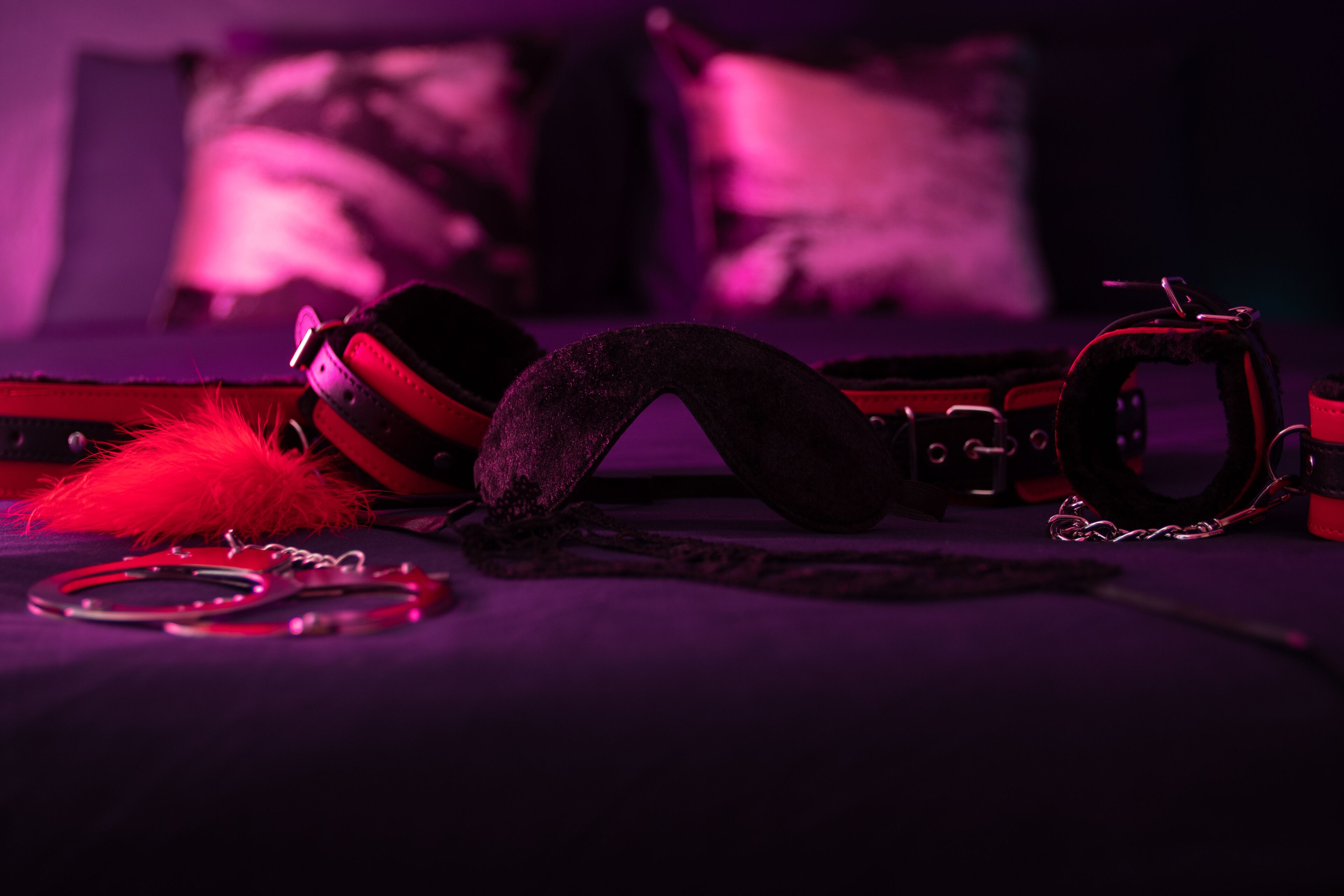 A bed with a black and red harness, a red feather, and handcuffs on a dark surface.