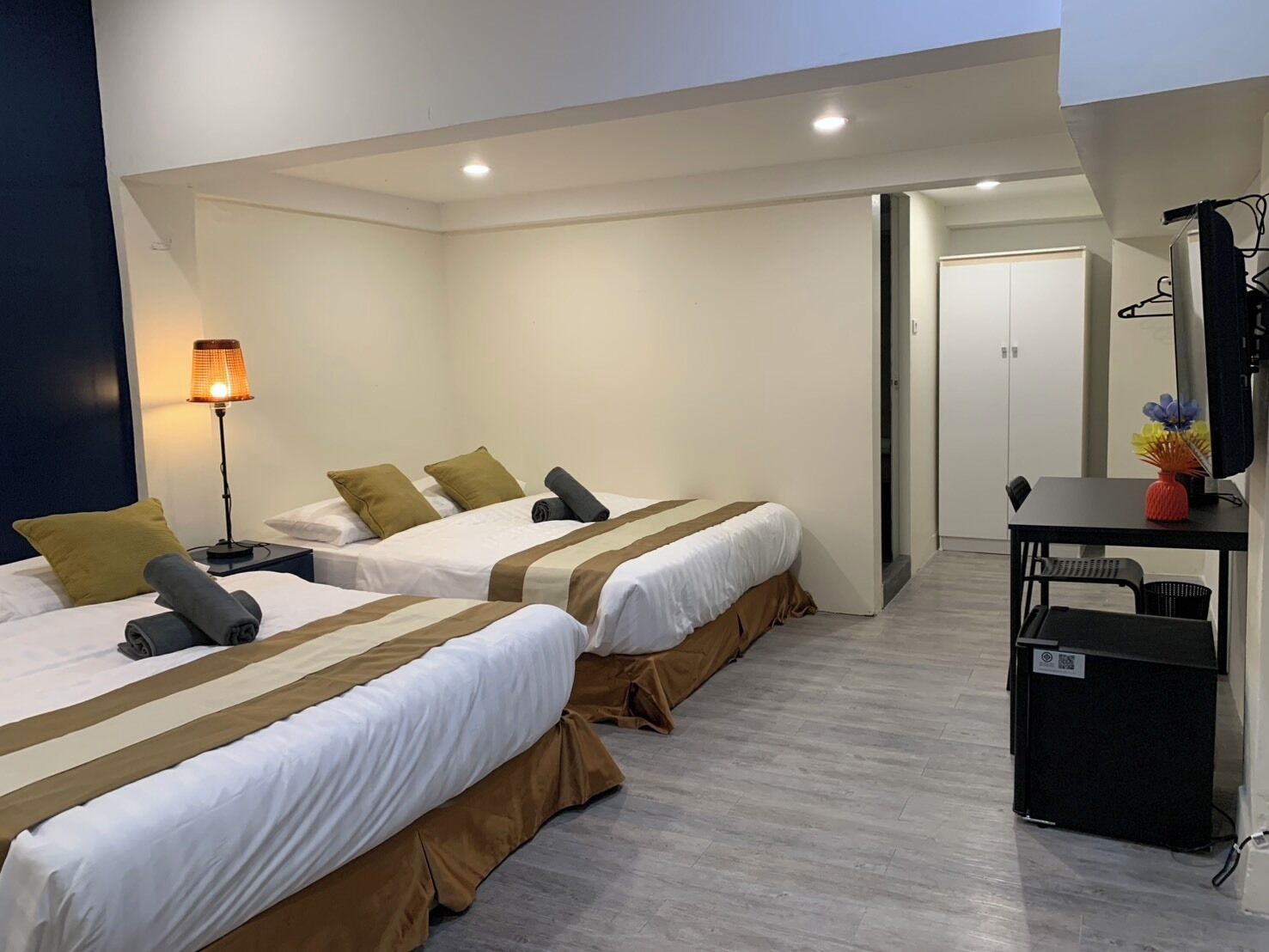 A hotel room with two beds, a desk, a lamp, and a small table with a vase.