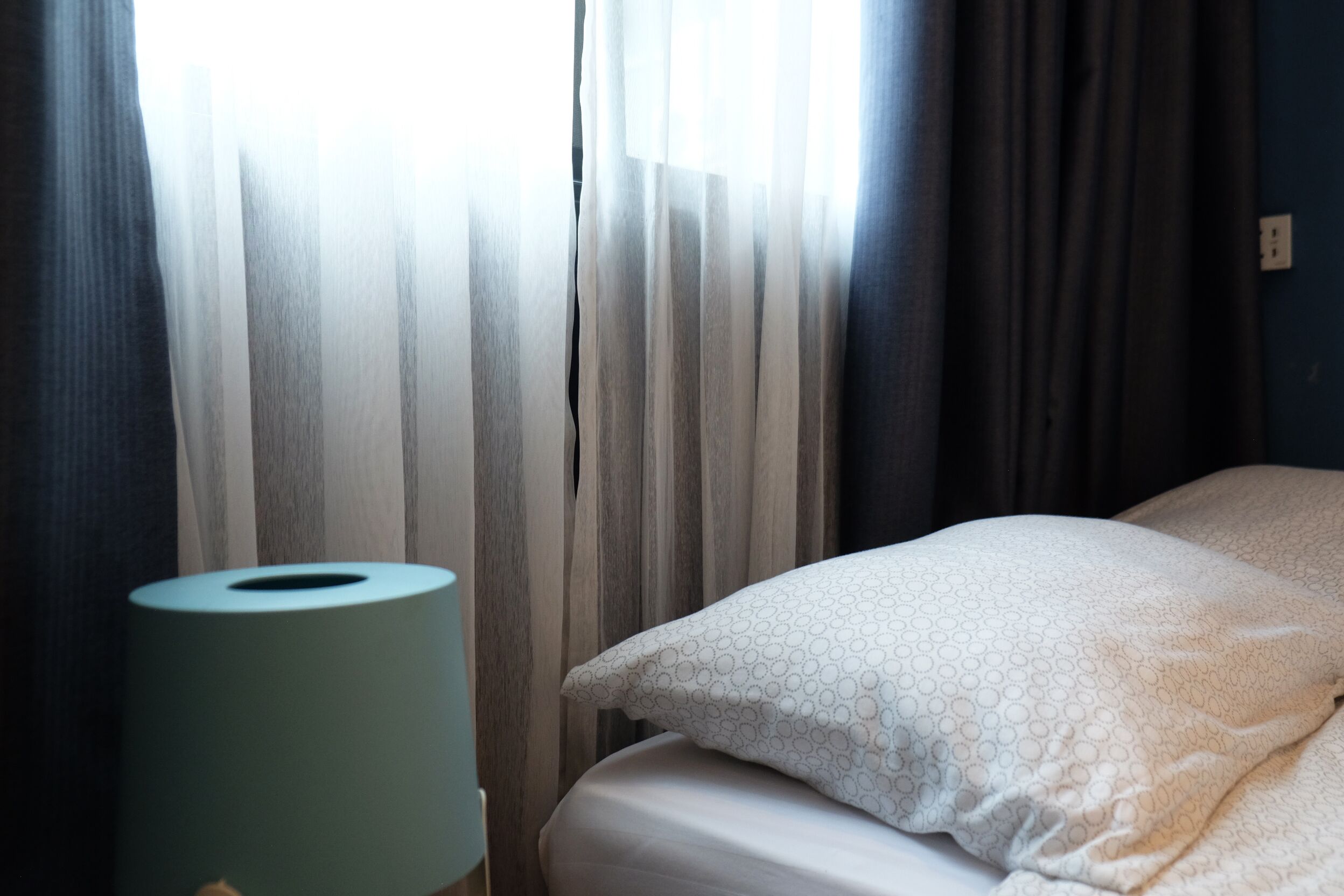 Una habitación de hotel con una cama, un almohadón, un rollo de papel higiénico y una ventana con cortinas.
