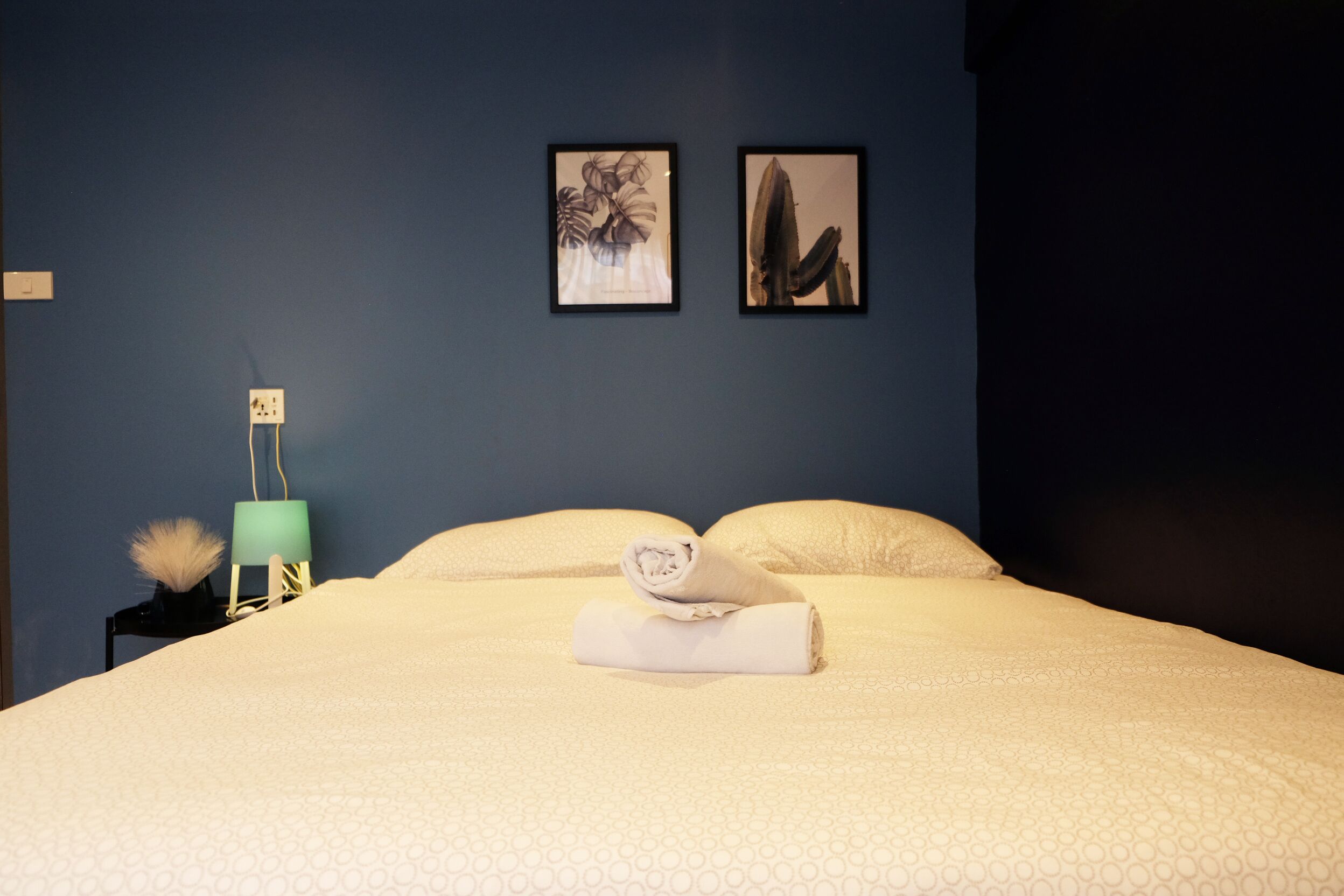 A neatly made bed with a rolled towel, a teal lamp, and two framed artworks on the wall.